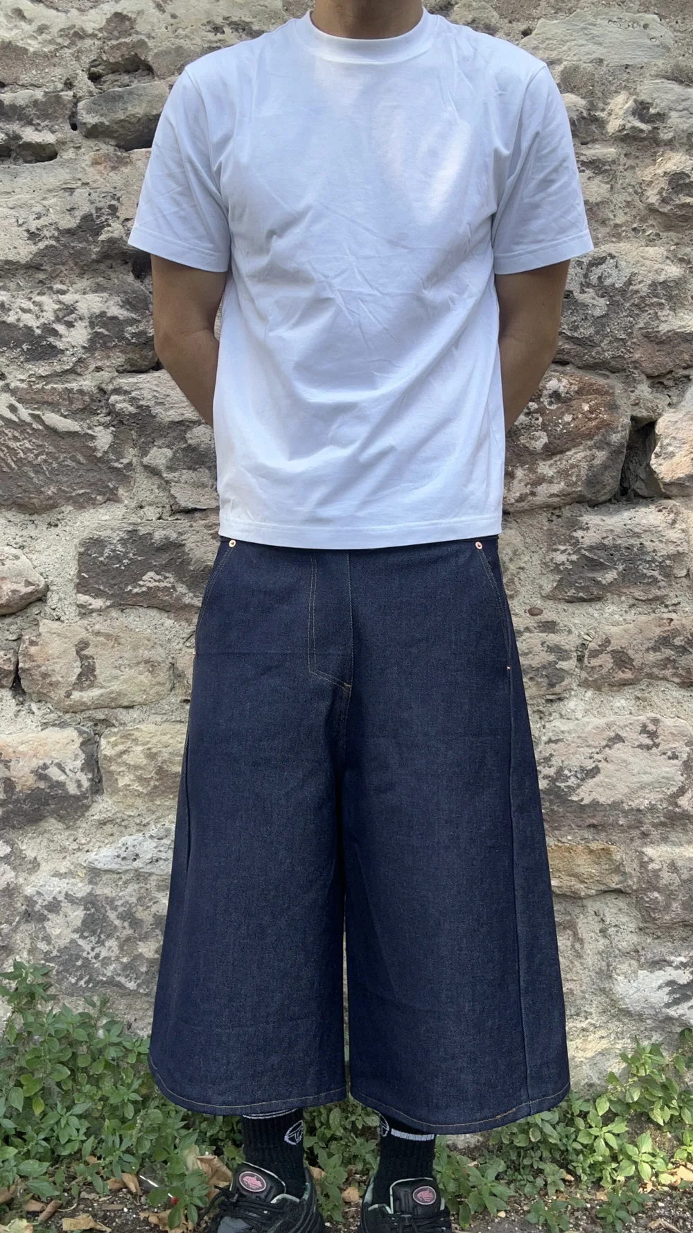 Pantalon en denim brut porté de face avec un tee-shirt blanc devant un mur en pierre